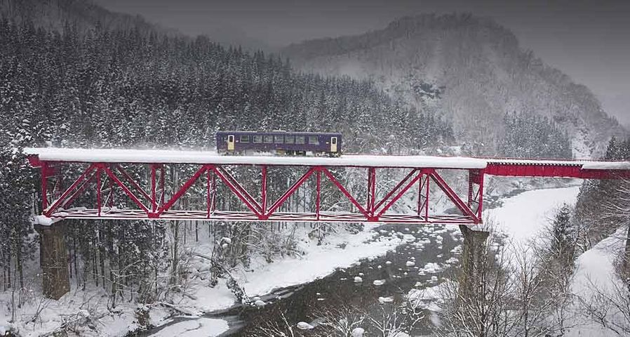 Ein Zug fährt über eine eiserne Brücke in verschneiter Landschaft, Region Tohoku, Japan – Japan Travel Bureau/Photolibrary ©