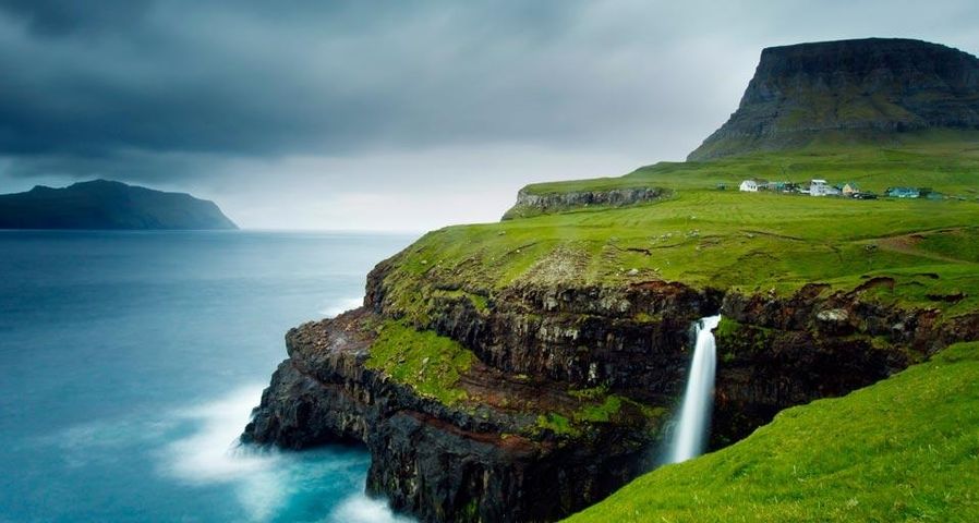 Village of Gásadalur below Heinanova mountain, with waterfall cascading over cliff into the Atlantic Ocean, Faroe Islands