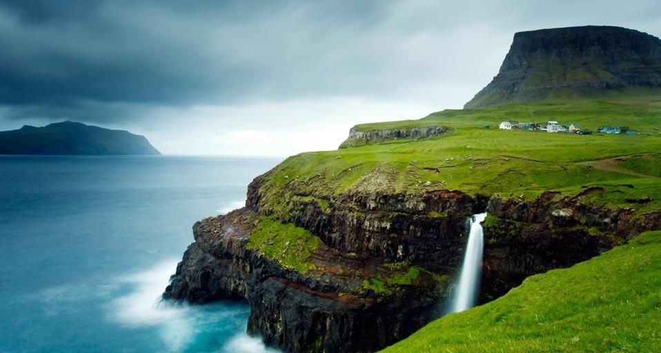Village of Gásadalur below Heinanova mountain, with waterfall cascading over cliff into the Atlantic Ocean, Vágar, Faroe Islands