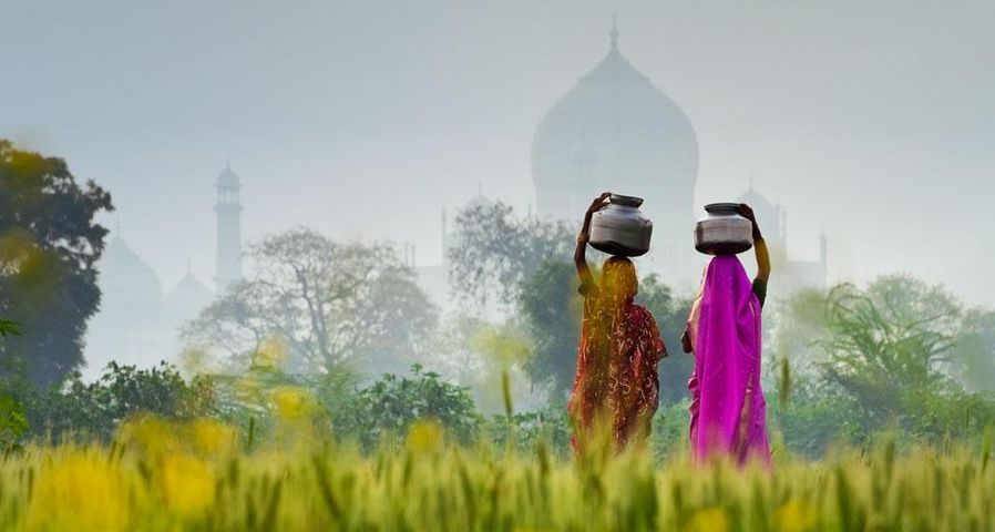 Zwei Frauen transportieren Wasserkrüge auf ihren Köpfen – im Hintergrund das Taj Mahal in Agra, Indien