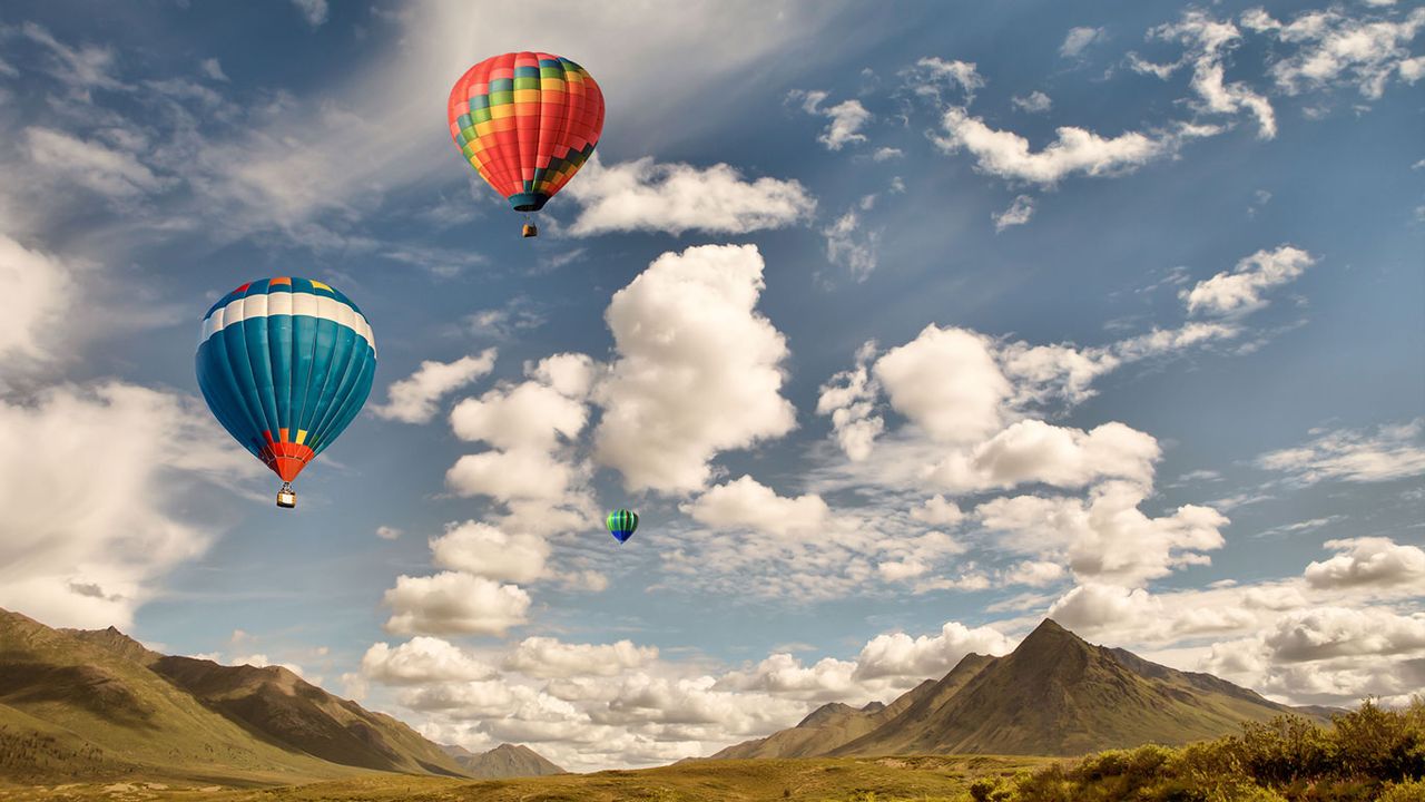 Three hot air balloons flying over mountain wilderness in Canada with puffy clouds and blue sky.