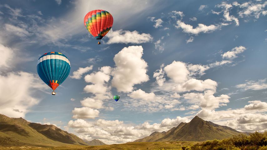 Three hot air balloons flying over mountain wilderness in Canada with puffy clouds and blue sky.